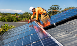 Técnico instalando paneles solares en el techo de una vivienda, con herramientas de seguridad y cielo despejado al fondo.